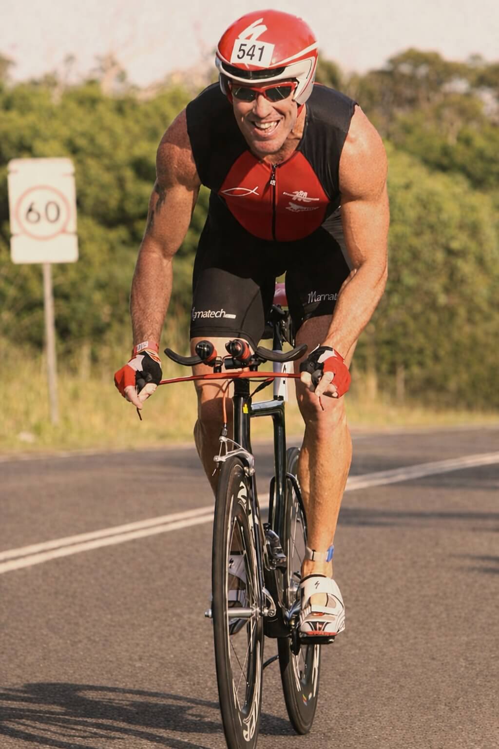 Athlete in red and black cycling gear riding a bicycle on a road with trees in the background.